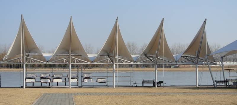 Row of peaked fabric canopies on steel masts beside the water, with boats stored on racks beneath
